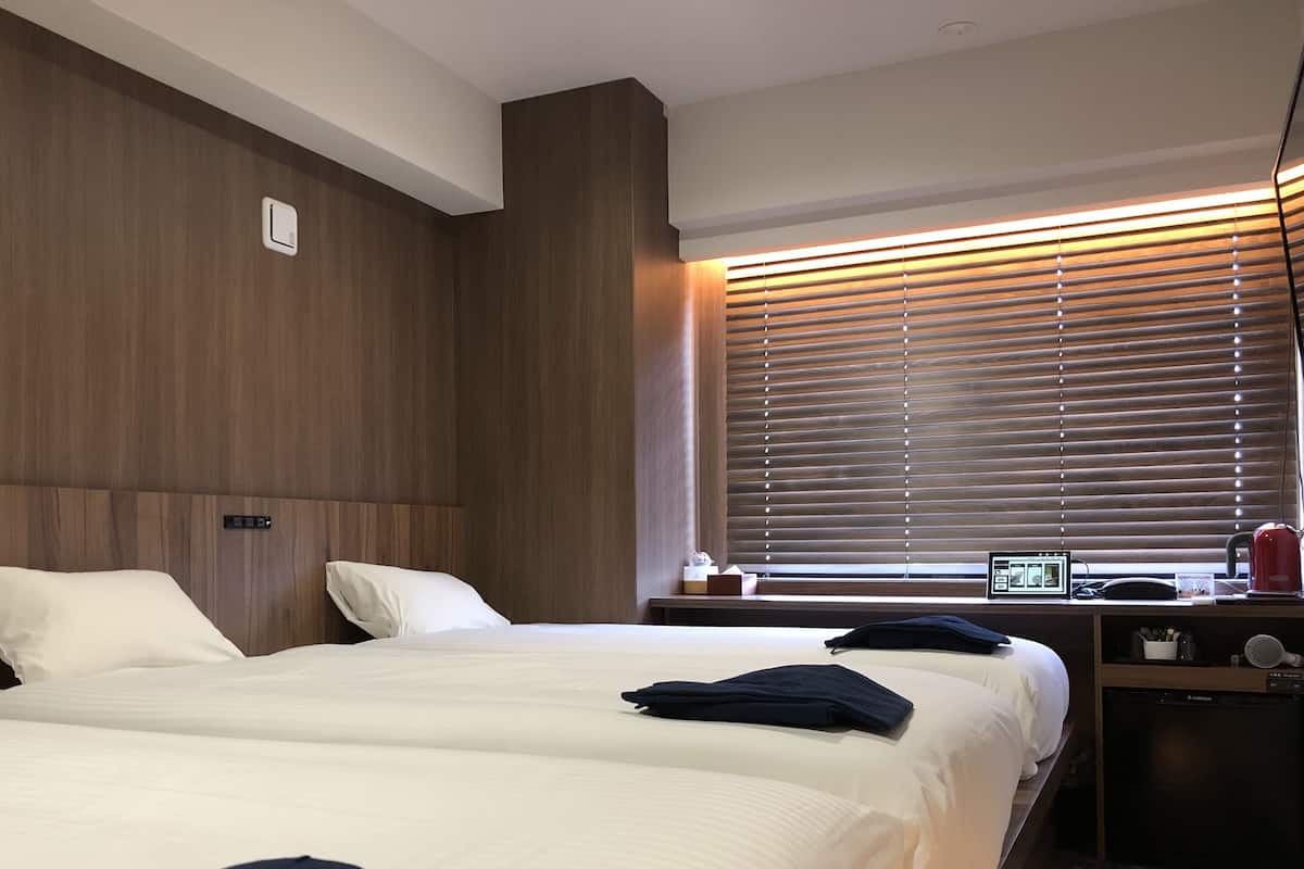 A hotel room with two beds, wooden paneling on the walls, a window with blinds, and a small table with a lamp and a digital device.