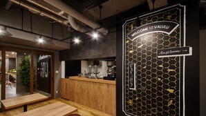Un café moderno con mesas y taburetes de madera, una barra con equipo para preparar café, y una pared con un diseño hexagonal y un letrero que dice 'Welcome to Valulu'.