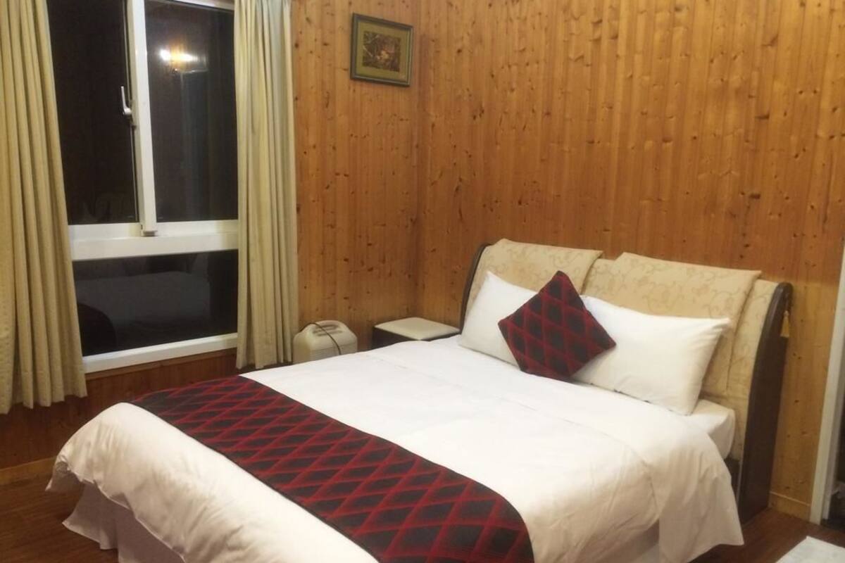 Una habitación de hotel con pared de paneles de madera, una cama grande con colcha estampada en rojo y negro, una mesita de noche y una ventana con cortinas.