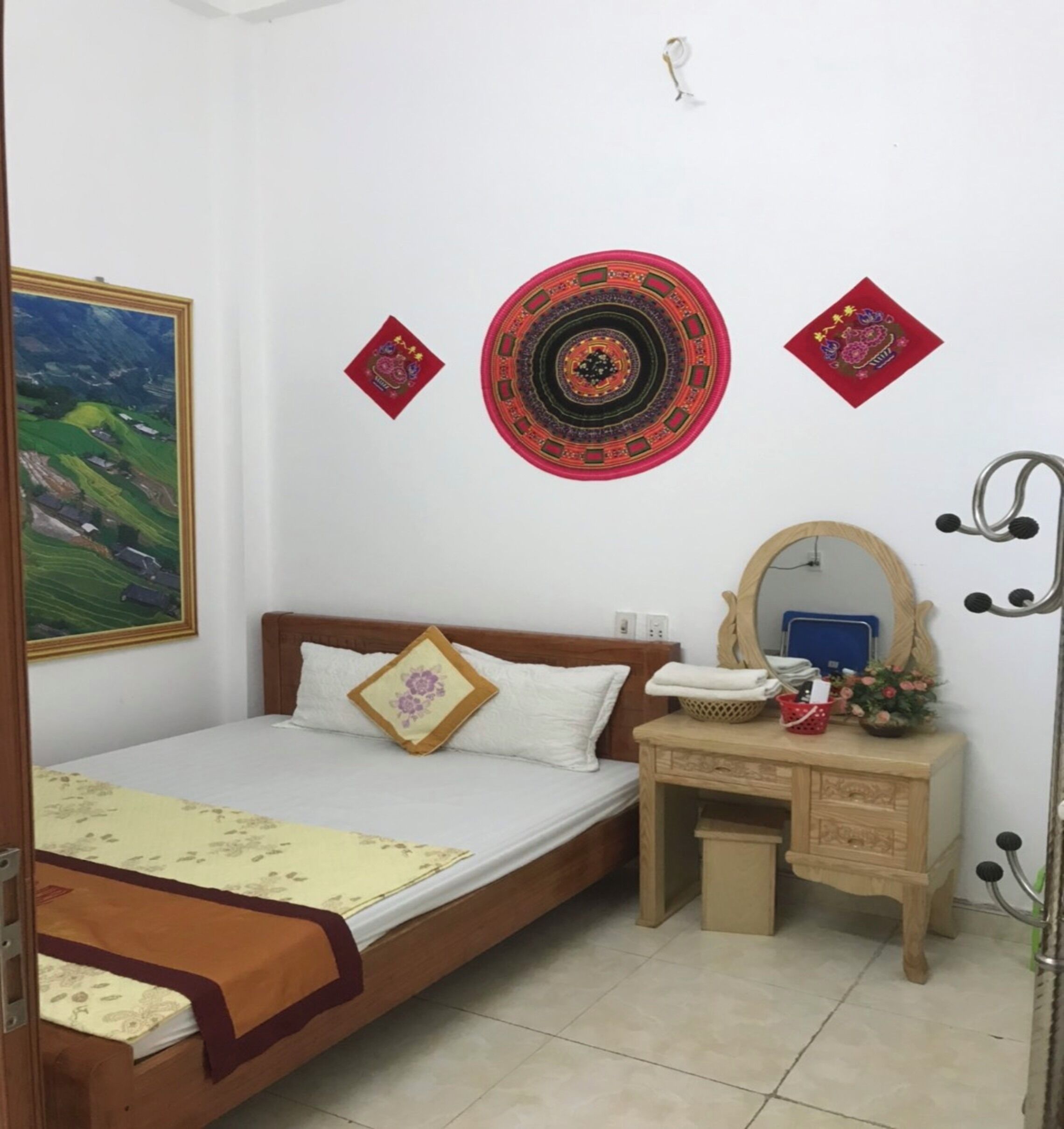 Ein Schlafzimmer mit Bett, Schminktisch, Spiegel und Wanddekorationen, darunter ein rundes, gemustertes Objekt und zwei rote, quadratische Elemente.