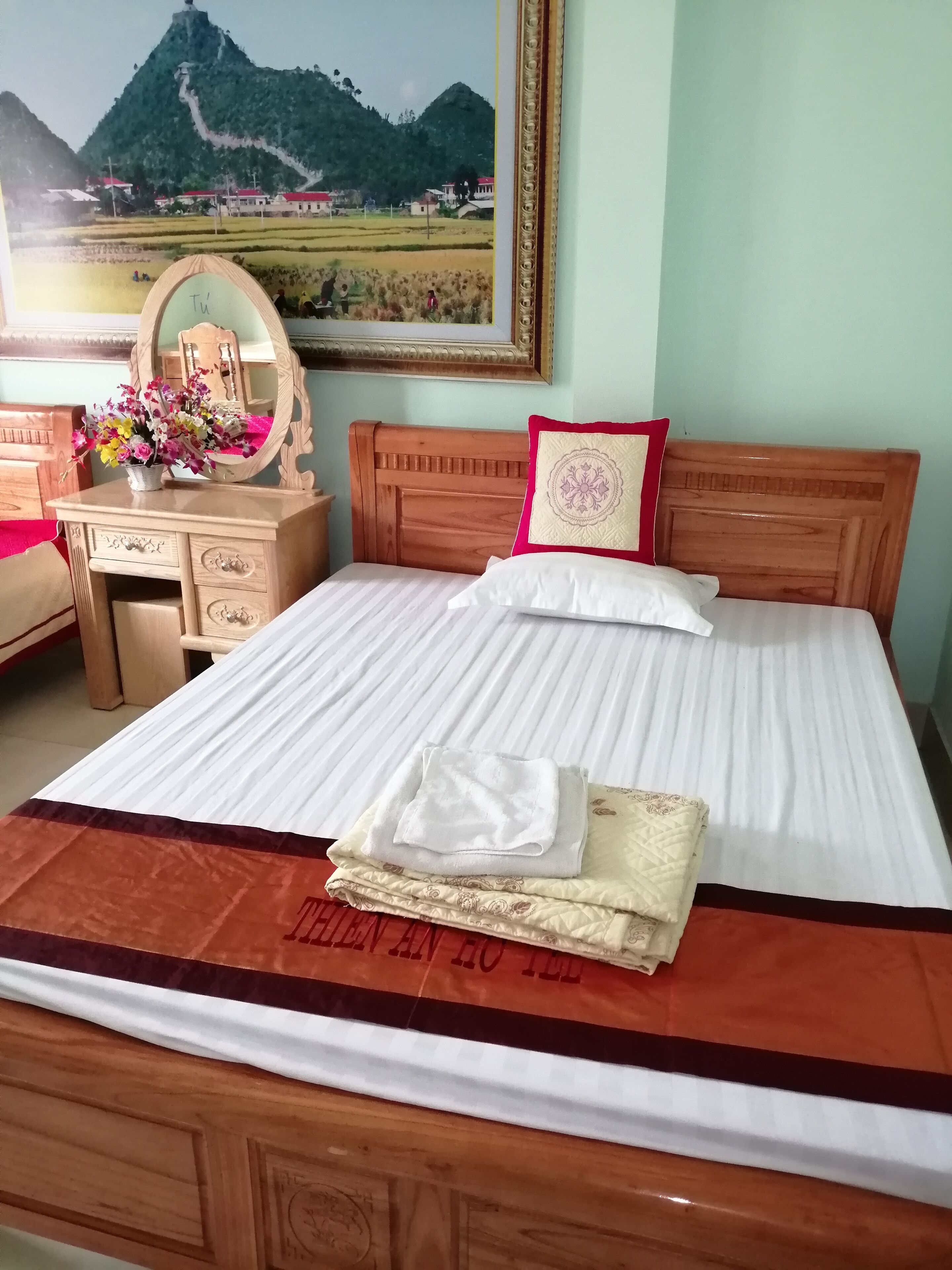 A neatly made bed with a white bedspread and a red pillow, a wooden dresser with a mirror and a vase of flowers, and a framed picture of a mountainous landscape on the wall.