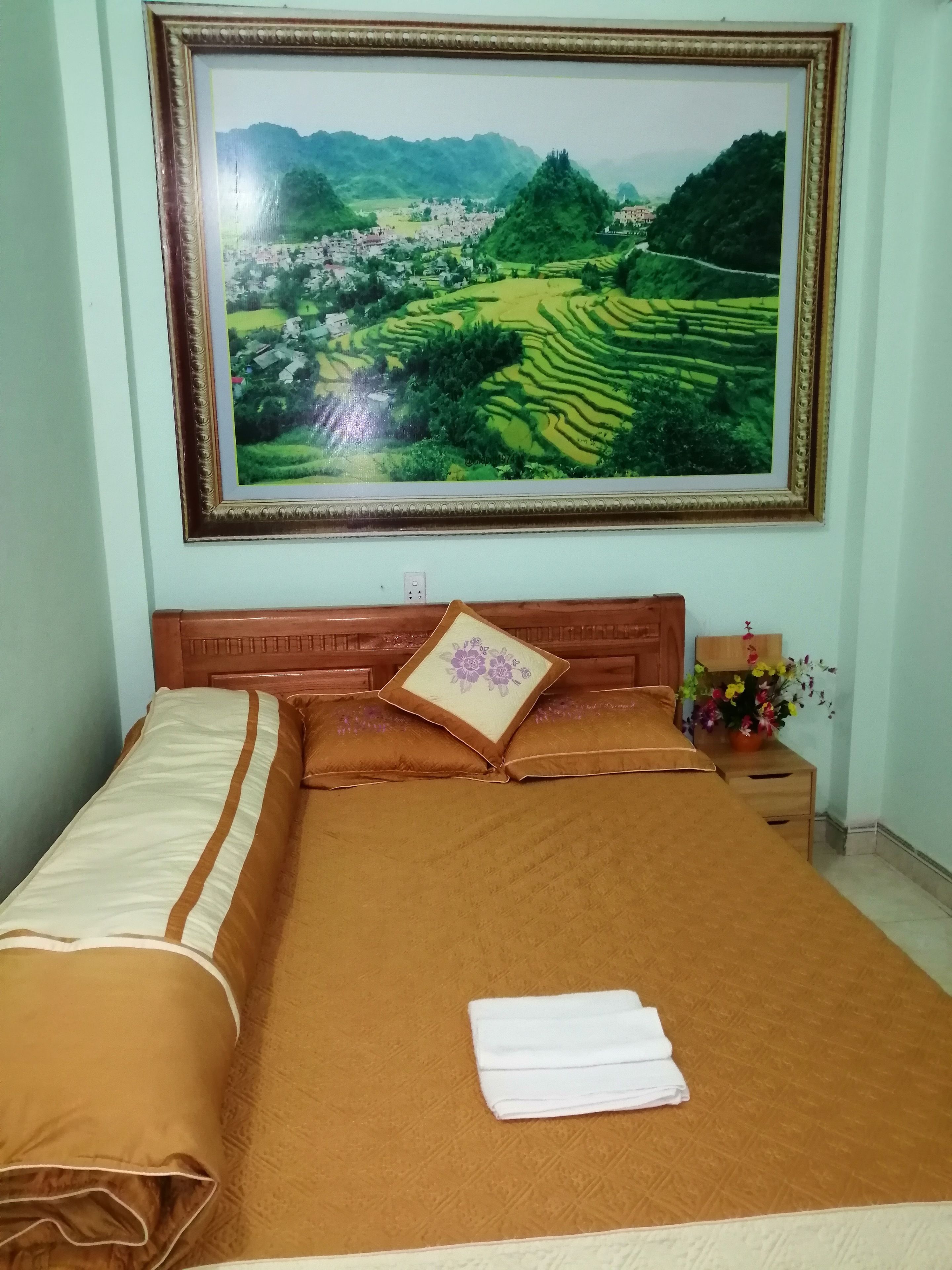 A neatly made bed with a brown bedspread and pillows, a framed landscape painting above it, and a small table with a vase of flowers beside the bed.