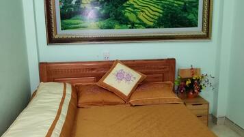 A neatly made bed with a brown bedspread and pillows, a framed landscape painting above it, and a small table with a vase of flowers beside the bed.