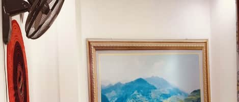 A hotel room with a large bed, a wooden dresser, a mirror, a wall-mounted fan, and a large framed landscape painting.