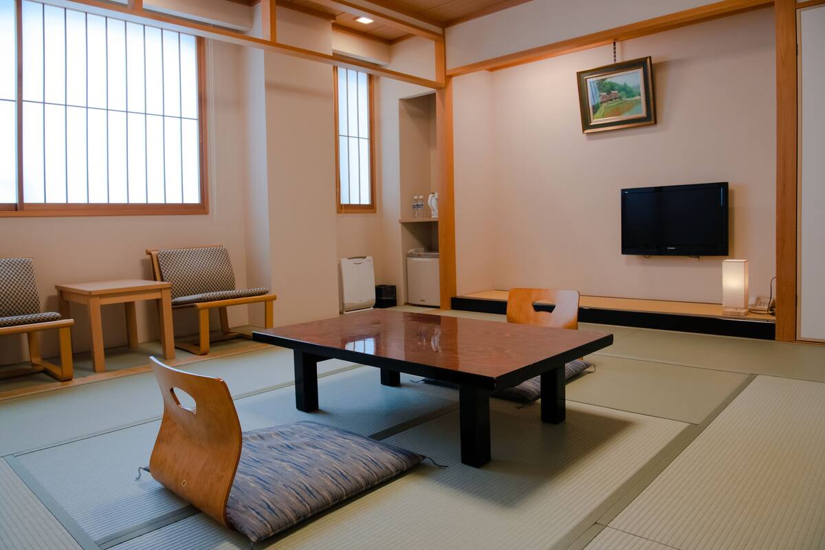Una habitación japonesa tradicional con suelo de tatami, una mesa baja, sillas, un televisor y un cuadro en la pared.