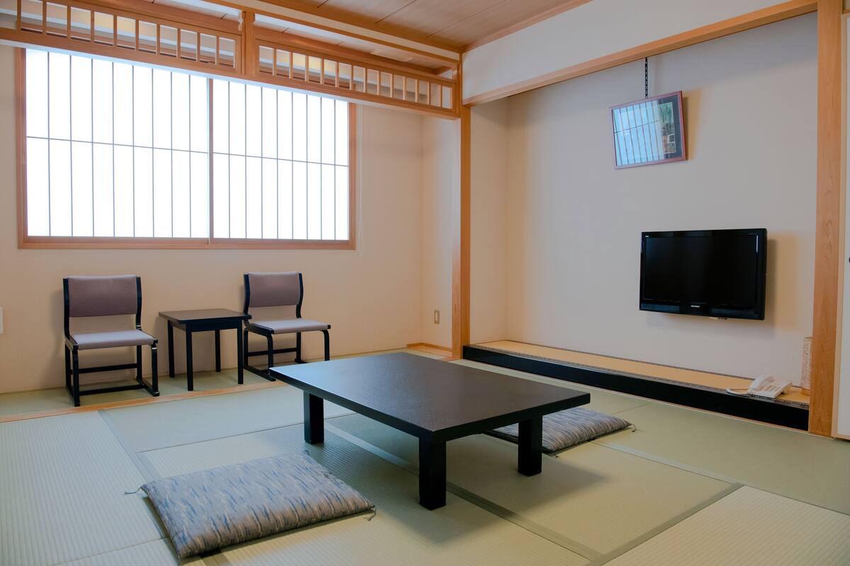 Una habitación japonesa tradicional con suelo de tatami, una mesa baja, dos sillas, un televisor y una puerta corredera.