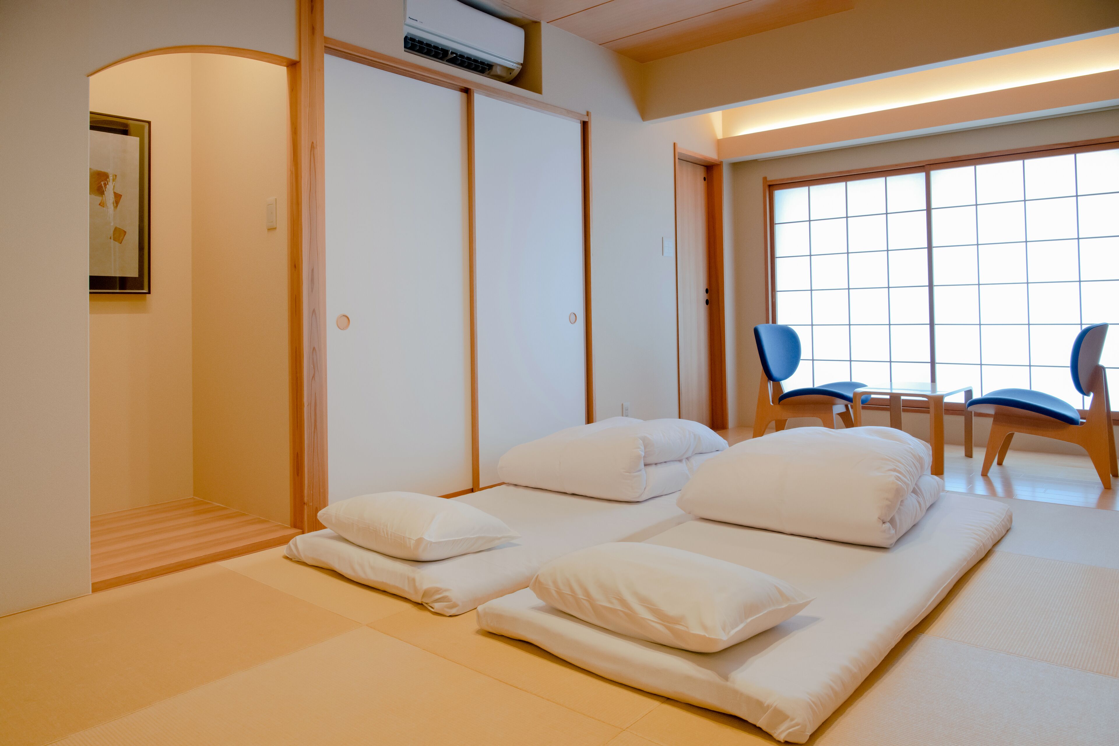 A room with tatami mats, a futon bed, a sliding door, and a view through a large window.