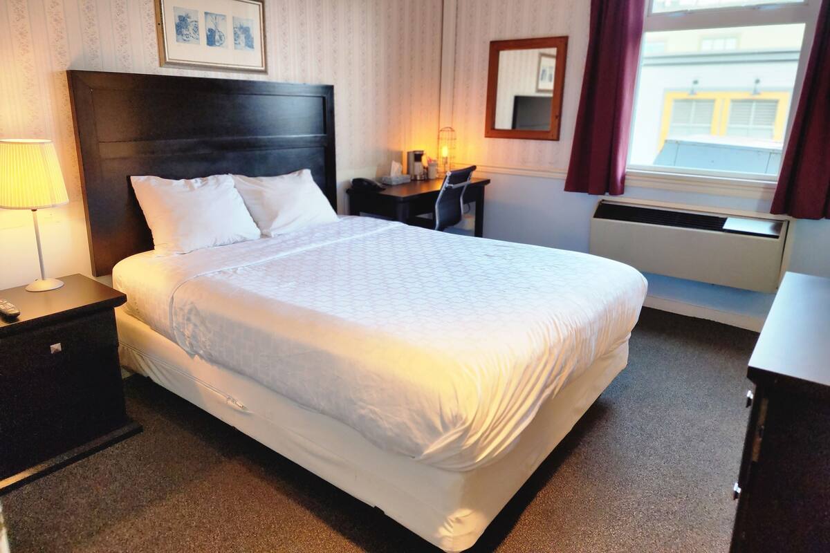 Une chambre d’hôtel avec un grand lit, une table de chevet avec une lampe, un bureau avec une chaise, une fenêtre avec des rideaux et un tableau encadré au mur.