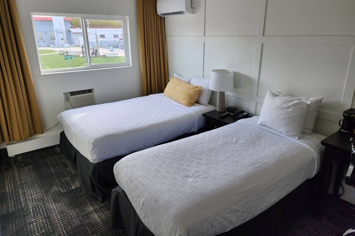 Une chambre d’hôtel avec deux lits, une fenêtre avec des rideaux, un appareil de climatisation, une table de chevet avec une lampe et un téléphone, et une vue sur l’extérieur.