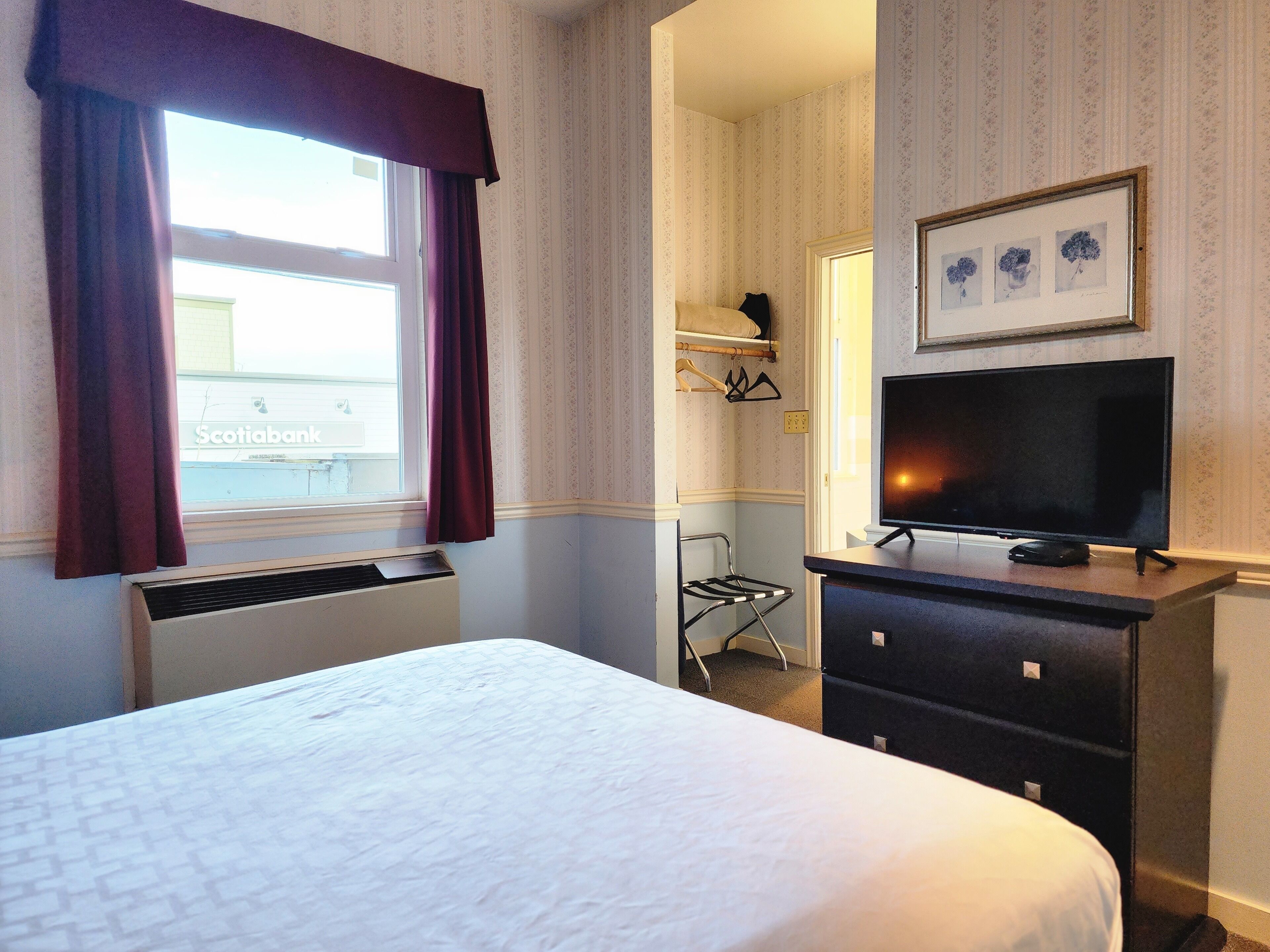 A hotel room with a bed, a flat-screen TV on a dresser, a window with curtains, and a wall-mounted shelf with a framed picture.