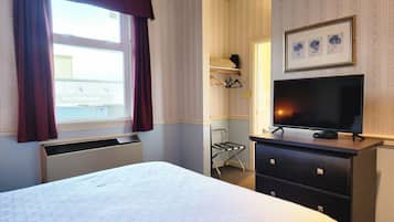 A hotel room with a bed, a flat-screen TV on a dresser, a window with curtains, and a wall-mounted shelf with a framed picture.