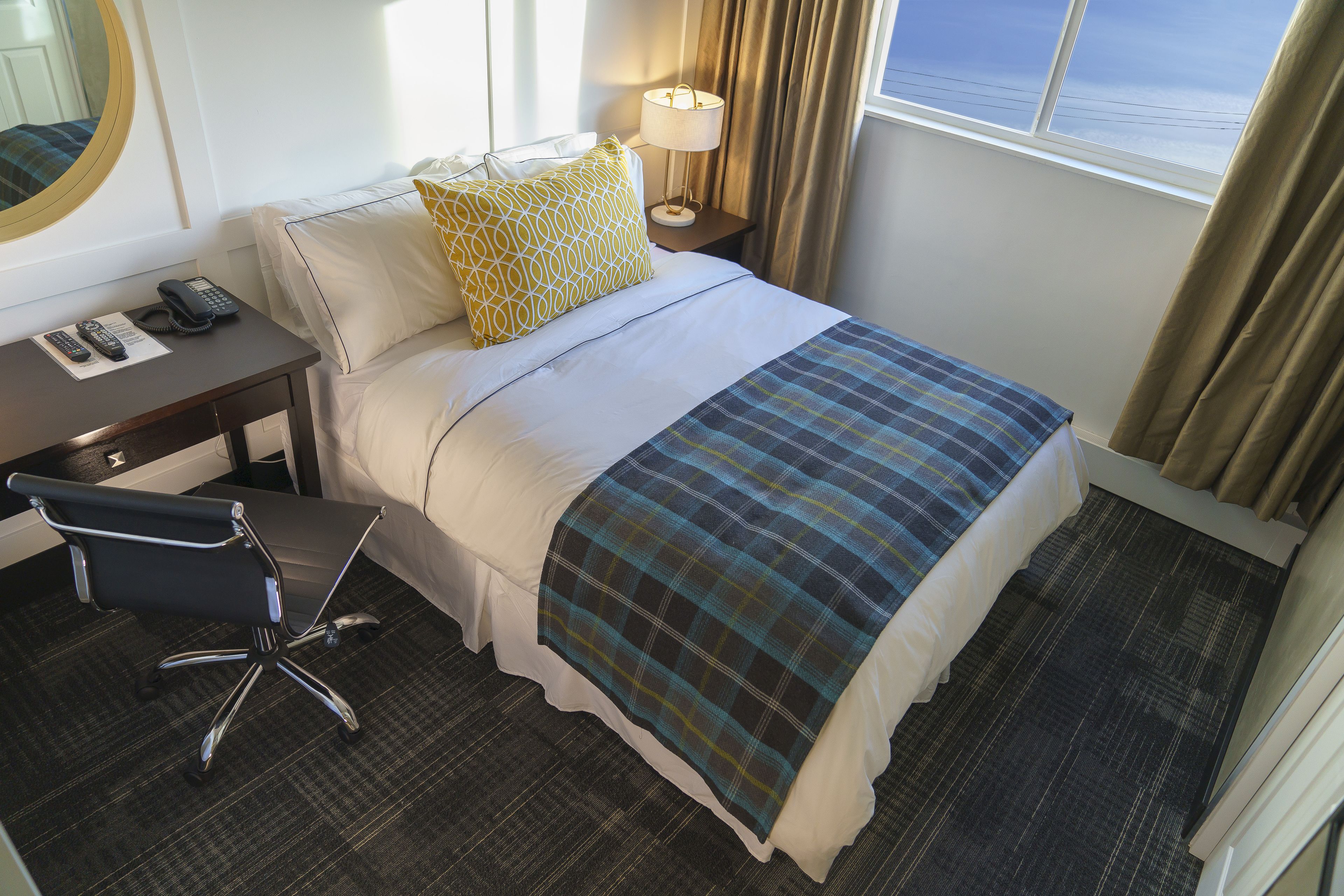A hotel room with a bed, a desk with a telephone, a chair, a lamp, and a window with curtains.