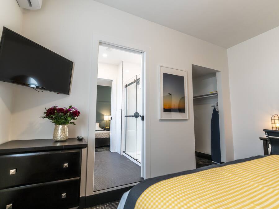 Una camera d'albergo con TV a schermo piatto montata a parete, un letto con copripiumino a righe gialle, un comò con cassetti, un vaso con fiori e una cornice con un quadro appeso al muro.
