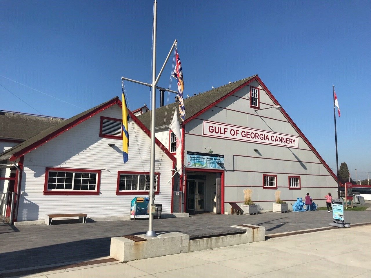 A building labeled "Gulf of Georgia Cannery" with a red and white exterior, a flagpole with multiple flags, and a clear blue sky.