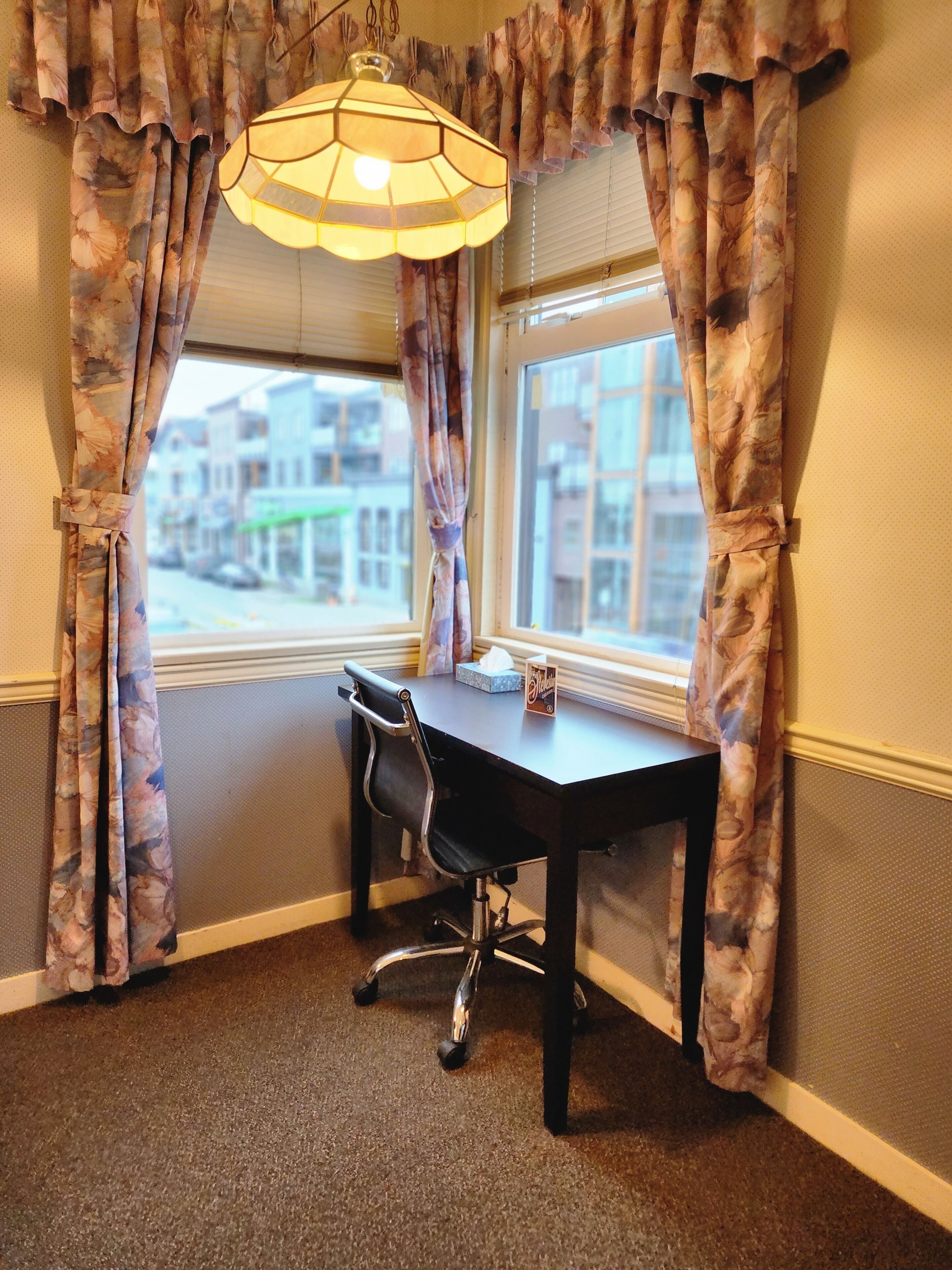 A corner office with a desk and chair, a window with curtains, and a hanging light fixture.