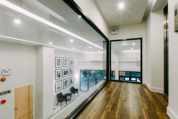 A modern hallway with wooden flooring, a glass wall with framed pictures, and an elevator.