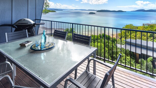 A balcony with a table, chairs, a grill, and a view of a large body of water and distant land. - Panorama 3 Hamilton Island 2 Bedroom Ocean View Near Marina With Golf Buggy (Whitsundays)