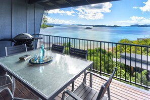 A balcony with a table, chairs, a grill, and a view of a large body of water and distant land. - Panorama 3 Hamilton Island 2 Bedroom Ocean View Near Marina With Golf Buggy (Whitsundays)
