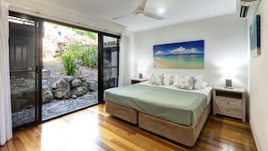A bedroom with a large bed, a ceiling fan, a nightstand, and a view of a garden through a sliding glass door. - Panorama 3 Hamilton Island 2 Bedroom Ocean View Near Marina With Golf Buggy (Whitsundays)