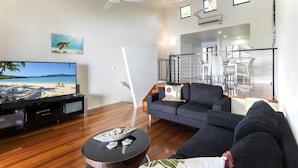 A modern living room with a flat-screen TV, a round coffee table, a dark sofa, and a staircase leading to an upper floor. - Panorama 3 Hamilton Island 2 Bedroom Ocean View Near Marina With Golf Buggy (Whitsundays)