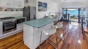 A modern kitchen with white cabinets, a central island with bar stools, a large window overlooking a scenic view, and a dining area with a view. - Panorama 3 Hamilton Island 2 Bedroom Ocean View Near Marina With Golf Buggy (Whitsundays)