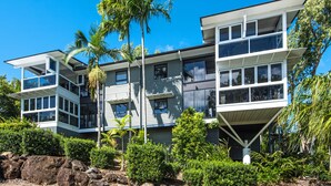 A modern two-story building with large windows and a flat roof, surrounded by lush greenery and palm trees. - Oasis 1 Hamilton Island 2 Bedroom Apartment In Central Location With Golf Buggy (Whitsundays)