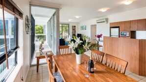 A spacious dining area with a wooden table, chairs, a vase of flowers, and a bottle of champagne. - Oasis 1 Hamilton Island 2 Bedroom Apartment In Central Location With Golf Buggy (Whitsundays)