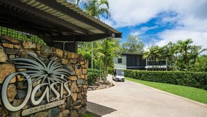 A stone wall with a sign reading "Oasis on Hamilton," a paved driveway, and a modern house in the background. - Oasis 1 Hamilton Island 2 Bedroom Apartment In Central Location With Golf Buggy (Whitsundays)