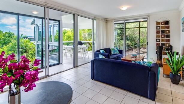 A modern living room with a blue sofa, a dining table, and a large sliding glass door leading to an outdoor patio. - Oasis 1 Hamilton Island 2 Bedroom Apartment In Central Location With Golf Buggy (Whitsundays)