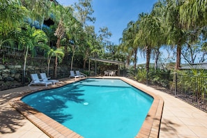 A swimming pool with a tiled edge, surrounded by palm trees and a covered seating area with chairs. - Shorelines 27 Hamilton Island 2 Bedroom Ocean View Near Marina With Golf Buggy (Whitsundays)