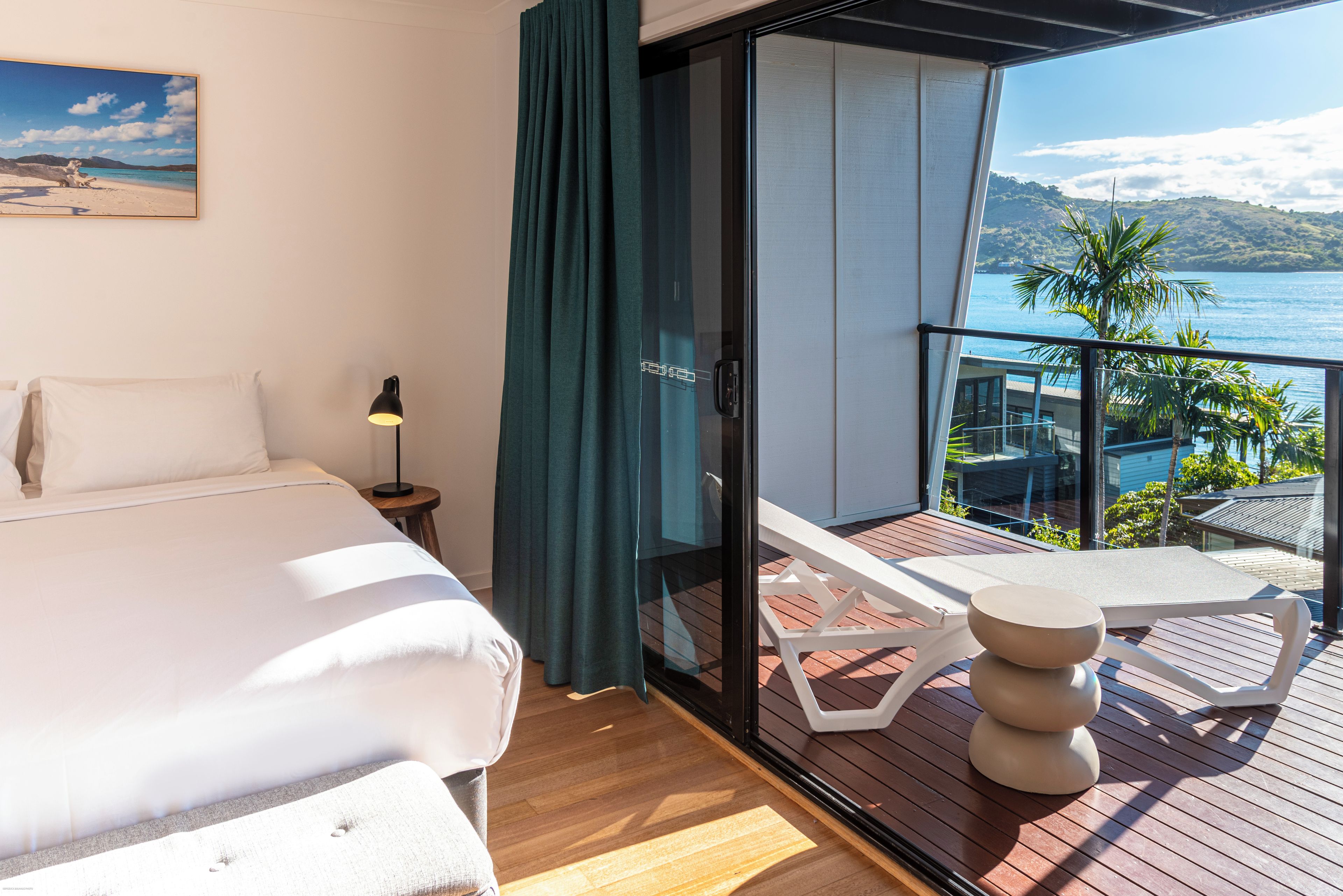 Una habitación de hotel con cama, mesita de noche, lámpara, balcón con vistas al mar y una mesa con un bol.