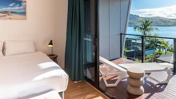 Una habitación de hotel con cama, mesita de noche, lámpara, balcón con vistas al mar y una mesa con un bol.