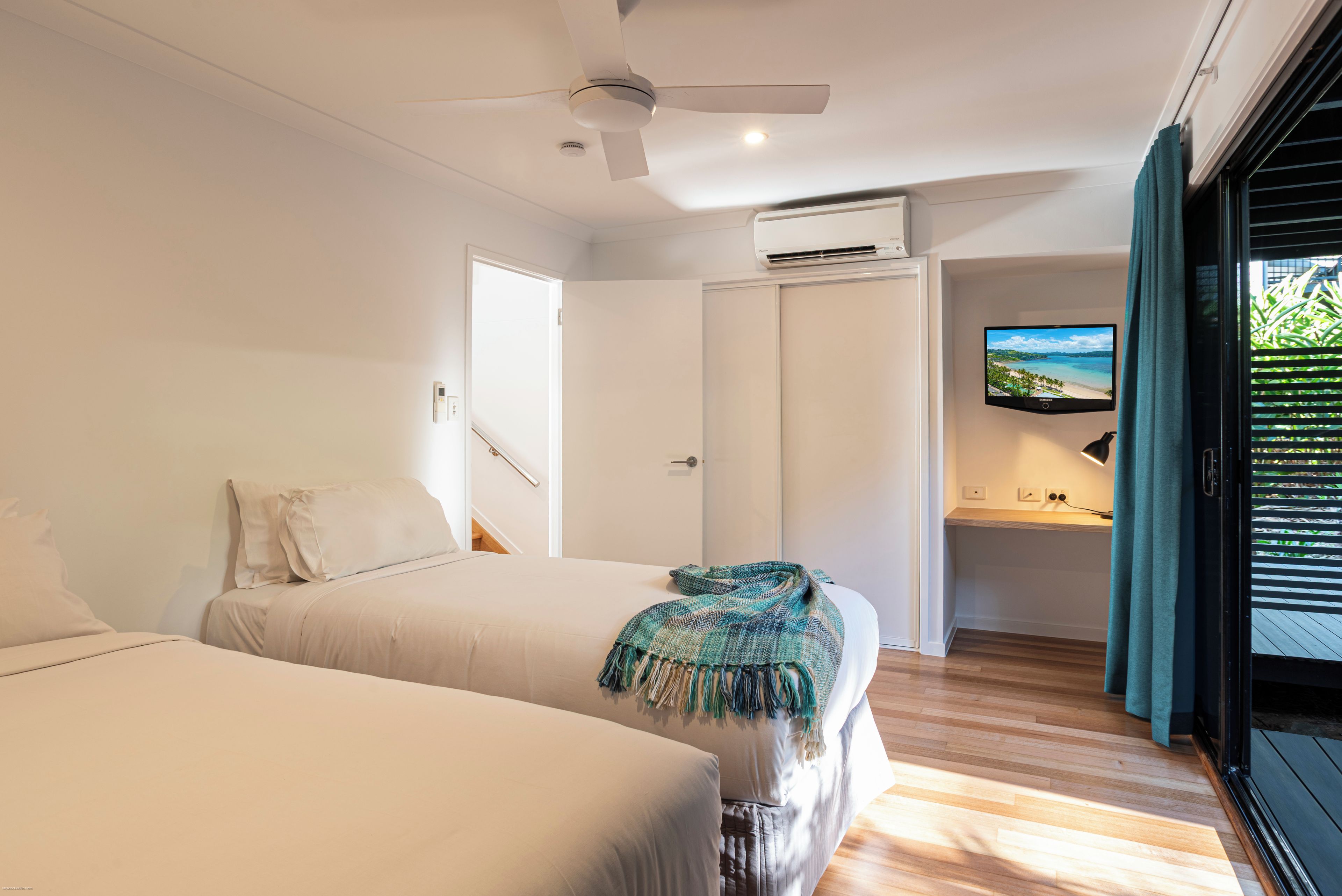 A bedroom with two beds, a ceiling fan, an air conditioning unit, a television, and a view of the outdoors through a sliding door.