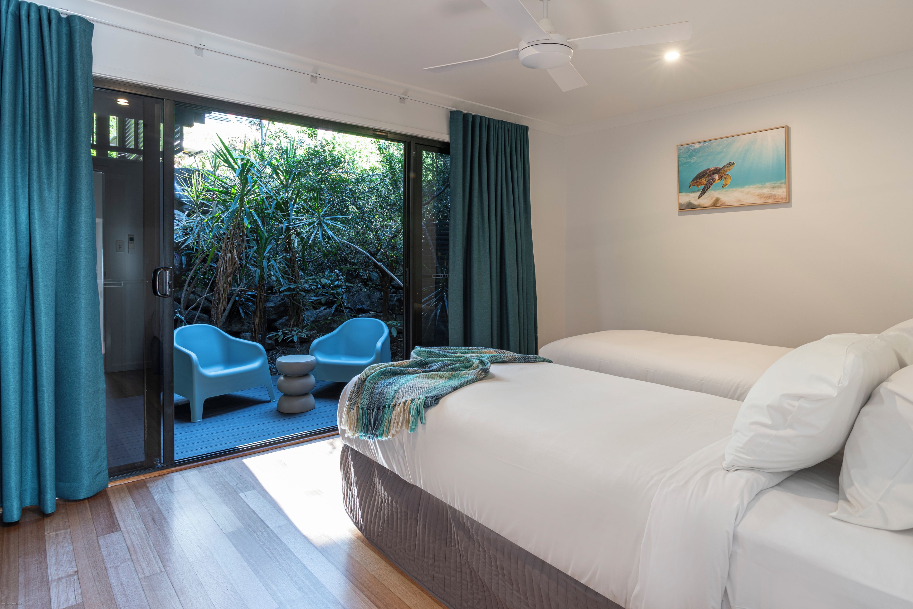 A bedroom with a bed, two chairs, a painting, and a view of greenery through a glass door.