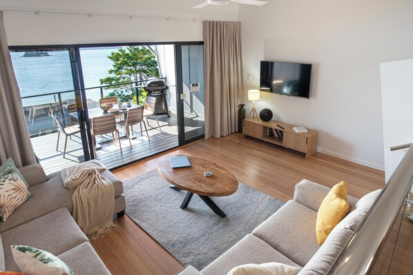 A modern living room with a sofa, a coffee table, a TV, and a balcony with a view of the sea. - Shorelines 27 Hamilton Island 2 Bedroom Ocean View Near Marina With Golf Buggy (Whitsundays)