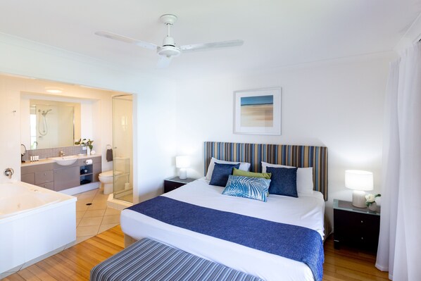 A hotel room with a large bed, a bedside table with a lamp, a ceiling fan, a bathroom with a shower and sink, and a framed picture on the wall.