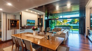 A spacious dining area with a wooden table and chairs, a kitchen with white cabinets, and a view of a lush garden through large windows.