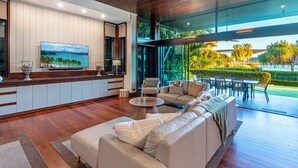 A modern living room with a large sofa, a coffee table, and a television. There is a view of a tropical landscape through large windows.