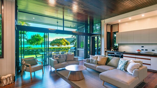 A modern living room with a large glass door leading to a balcony, a kitchen with white cabinets, and a wooden floor.