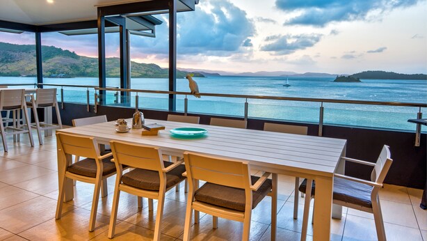 A dining area with a table and chairs, overlooking a body of water with distant land and a sunset. - Villa 16 The Edge Oceanfront Deluxe 3 Bedroom Near Marina With Golf Buggy (Whitsundays)
