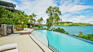 A modern pool area with lounge chairs, a stone wall, and a view of a lake. - Villa 16 The Edge Oceanfront Deluxe 3 Bedroom Near Marina With Golf Buggy (Whitsundays)
