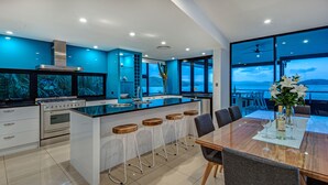 A modern kitchen with a central island, bar stools, and a dining area with a view of the sea. - Villa 16 The Edge Oceanfront Deluxe 3 Bedroom Near Marina With Golf Buggy (Whitsundays)