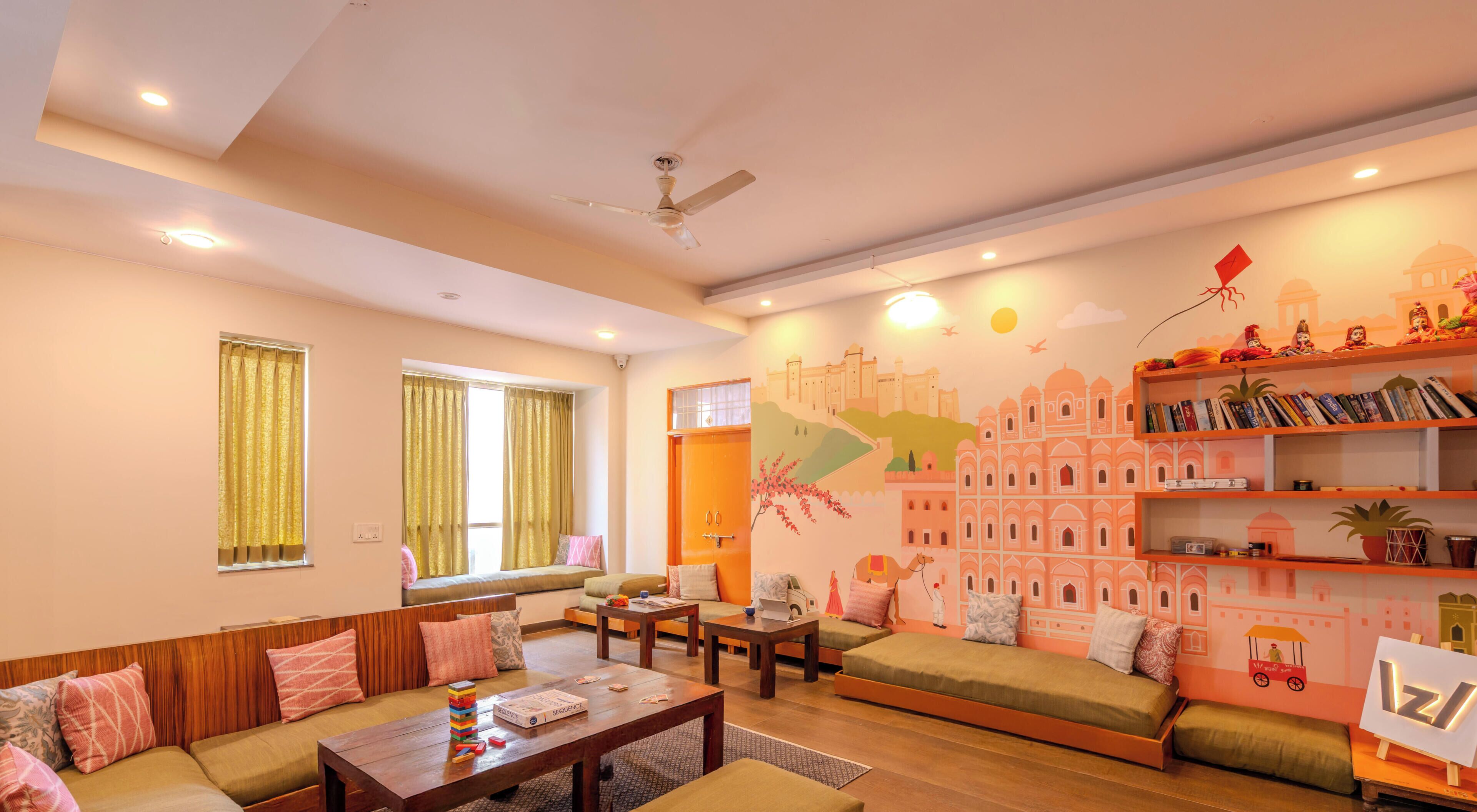 Una sala de estar con un juego de sofá, una mesa de centro con libros y un juego de mesa, una estantería con libros y objetos decorativos, y un mural de un paisaje urbano en la pared.