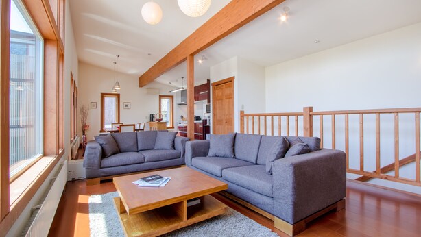 A spacious living room with a large grey sectional sofa, a wooden coffee table, and a view of the kitchen in the background. - Cocoa Niseko (Kutchan)