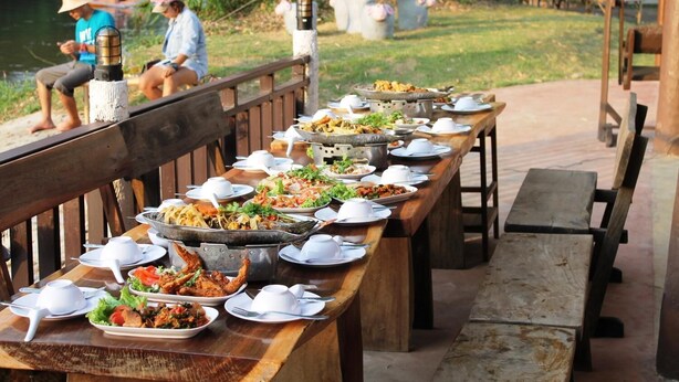 A long wooden table set with various dishes and bowls, likely for a communal meal, outdoors with a view of a body of water and people in the background.