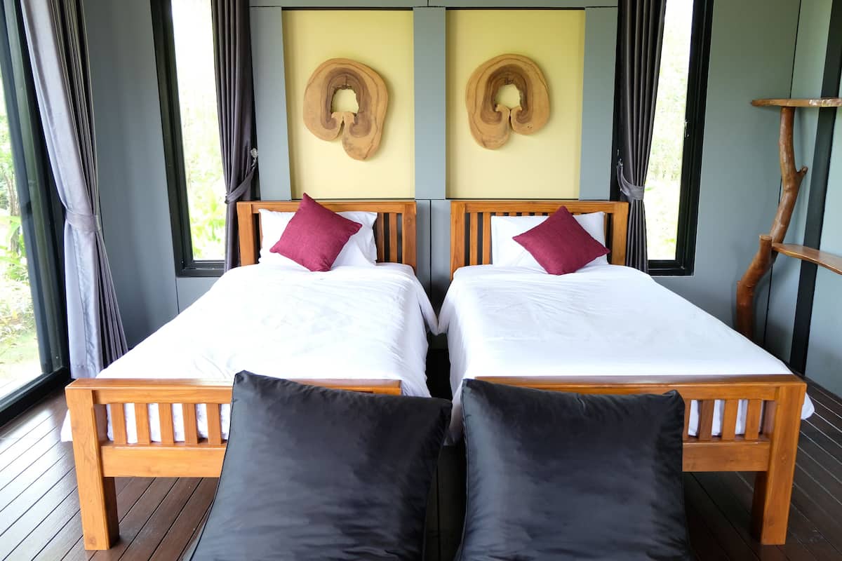 A double bed with white linens and two red pillows, a wooden headboard, and a view of greenery through a window.
