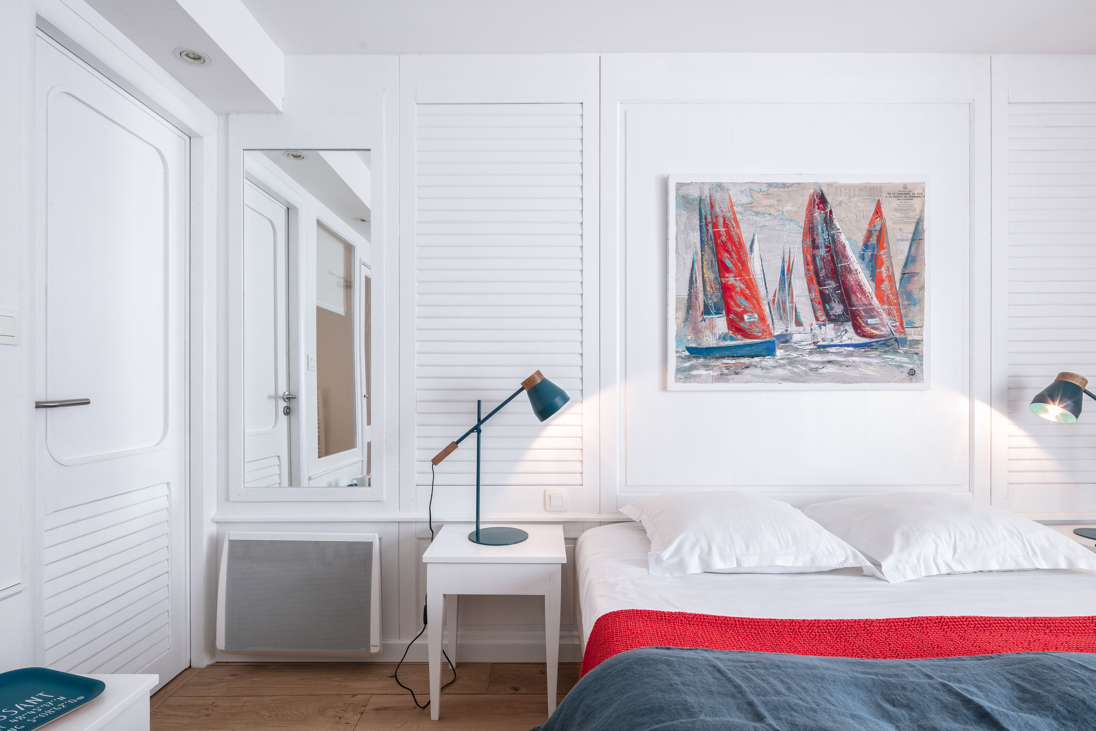 Un dormitorio con cama, mesita de noche, lámpara, un cuadro de veleros y ventana con persianas.