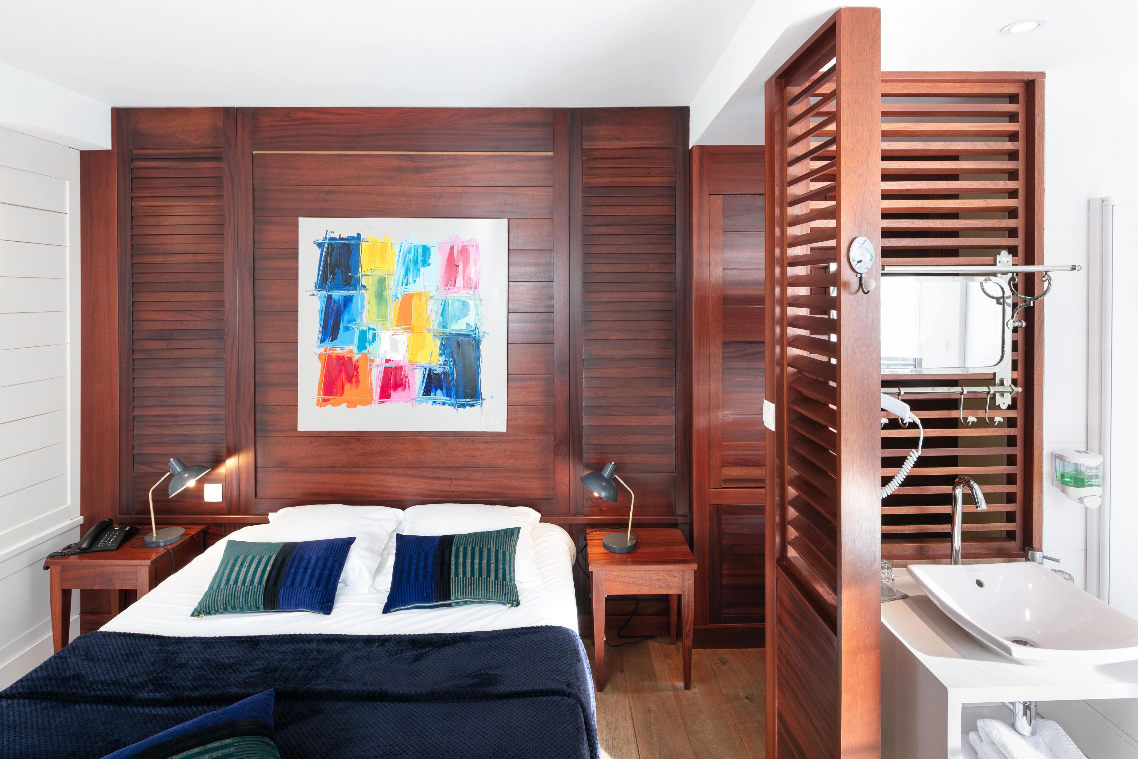 Una habitación de hotel con una cama grande, dos mesitas de noche con lámparas, un cabecero de madera, un cuadro abstracto colorido y un baño con lavamanos y espejo.