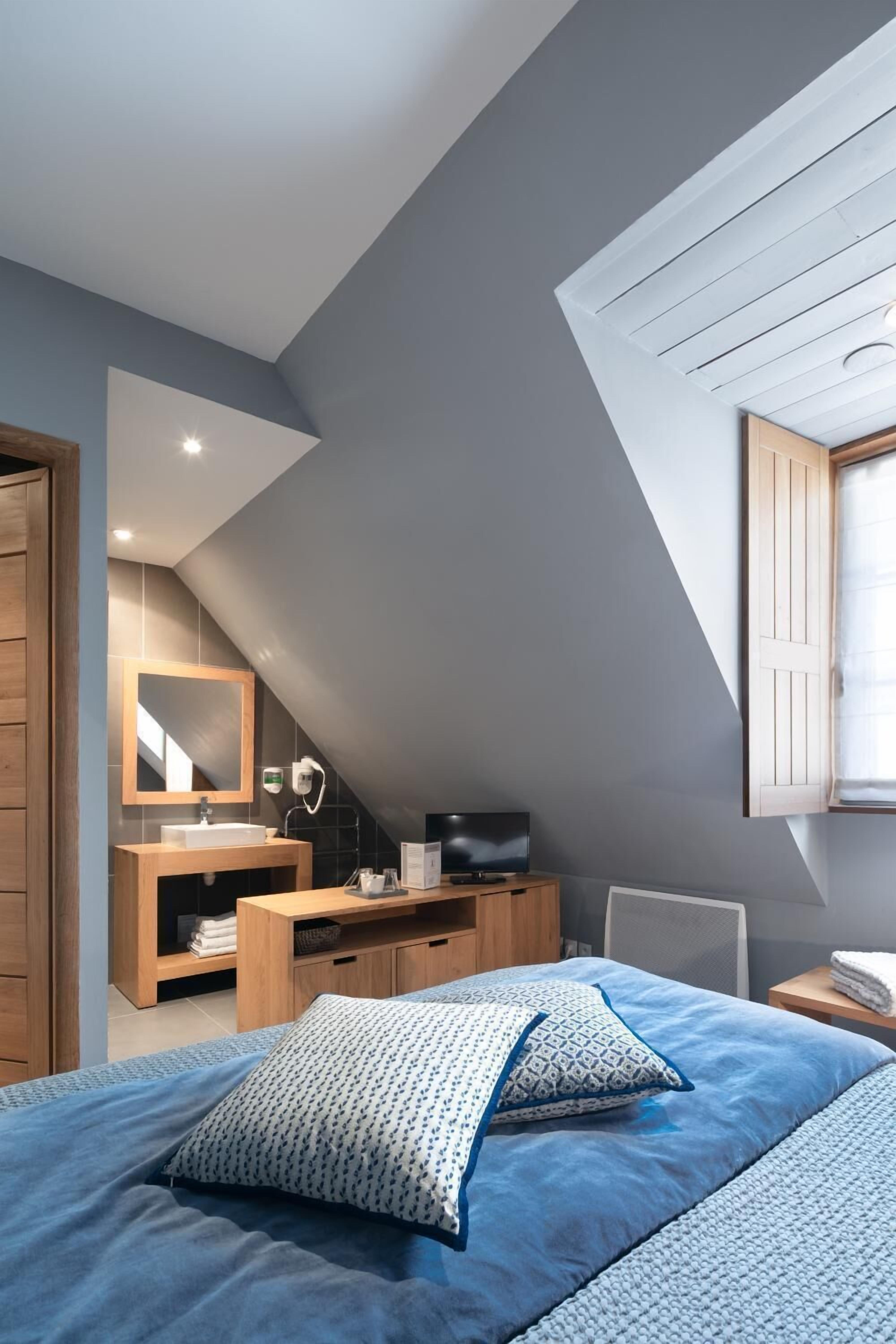A bedroom with a sloped ceiling, a bed with blue bedding, a wooden nightstand, a TV, and a window with a view.