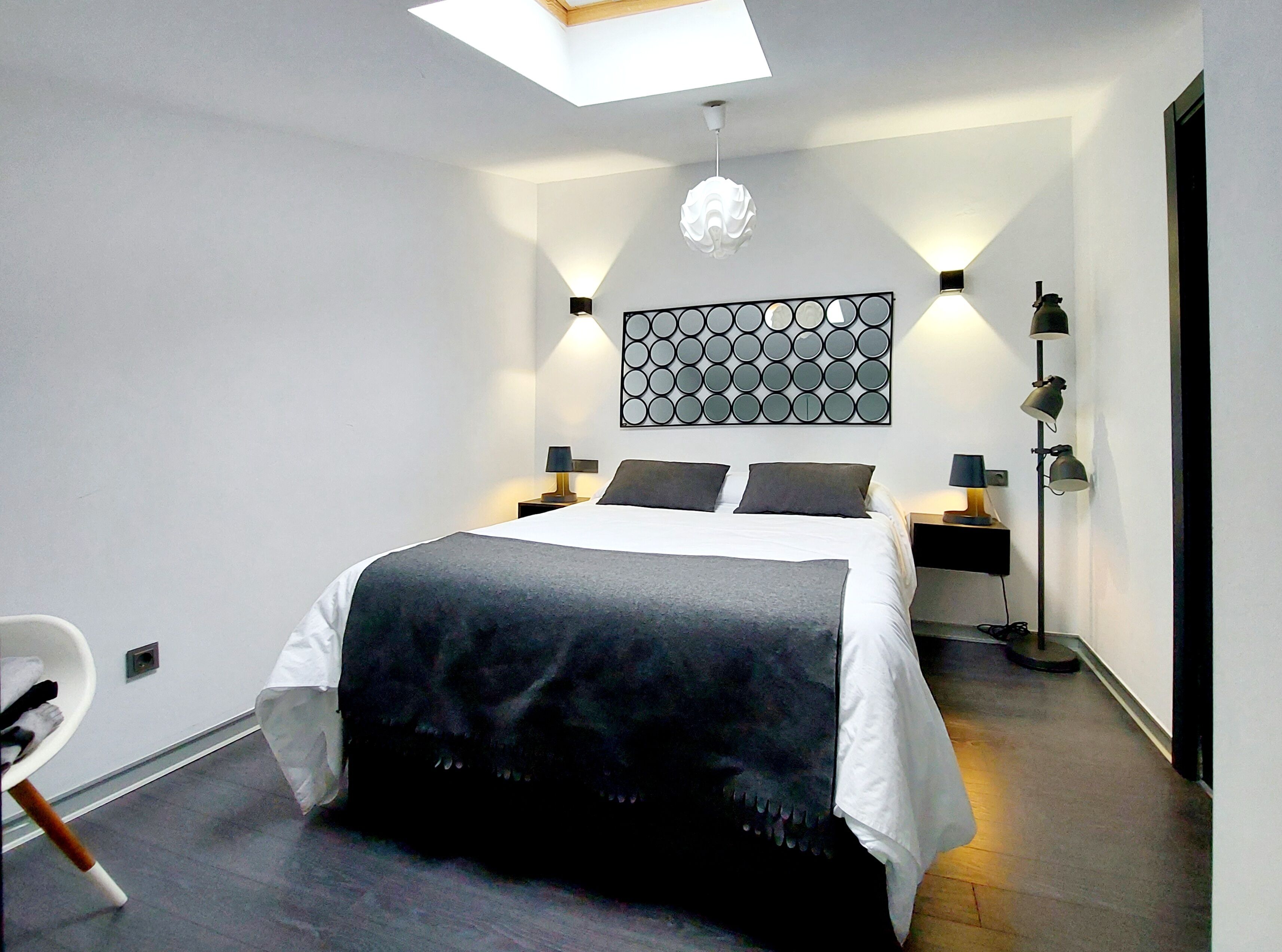 Un dormitorio moderno con tragaluz, cama con colcha blanco y negro, dos mesitas de noche con lámparas y una lámpara de pared.